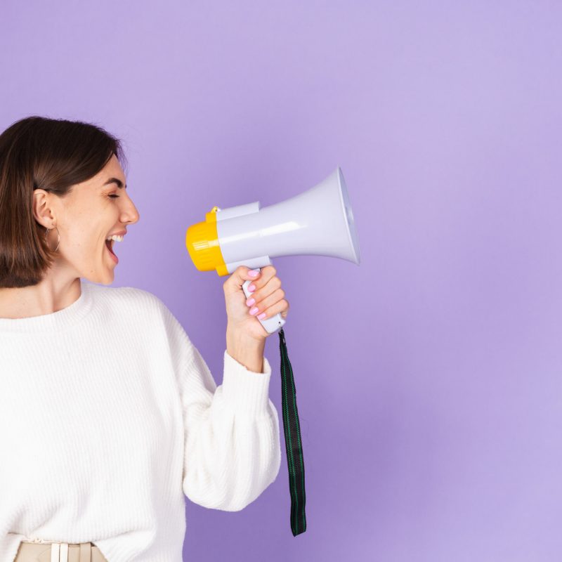 Young brunette in white casual sweater isolated on purple background happy screaming in megaphone copy space point left